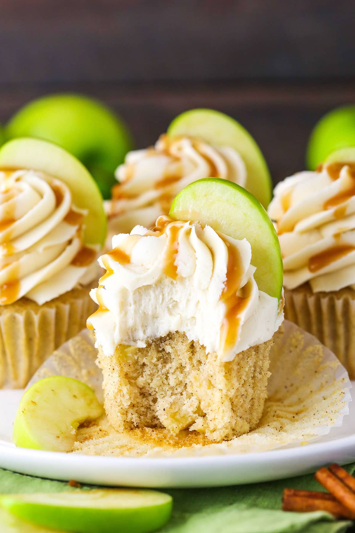 These Caramel Apple Cupcakes are made with a spiced apple cupcake and topped with caramel icing. They melt in your mouth and are such a delicious apple dessert idea! A Caramel Apple Cupcake with a bite taken out on a white plate with more Caramel Apple Cupcakes in the background