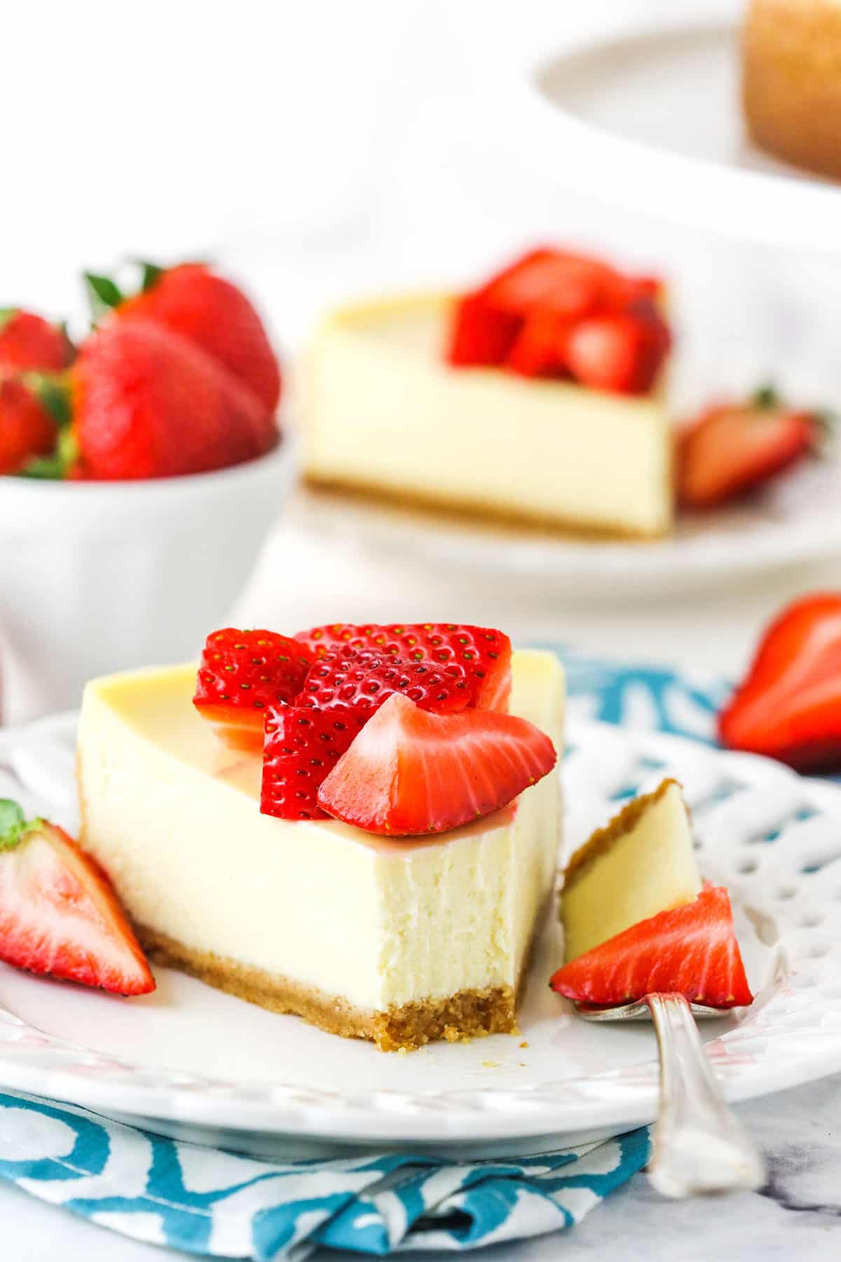 This easy Instant Pot Cheesecake recipe has classic flavors with graham cracker crust and strawberry topping! Perfect, creamy, no-fuss cheesecake! A slice of Instant Pot Cheesecake topped with cut strawberries with a bite taken out on a white plate with a fork