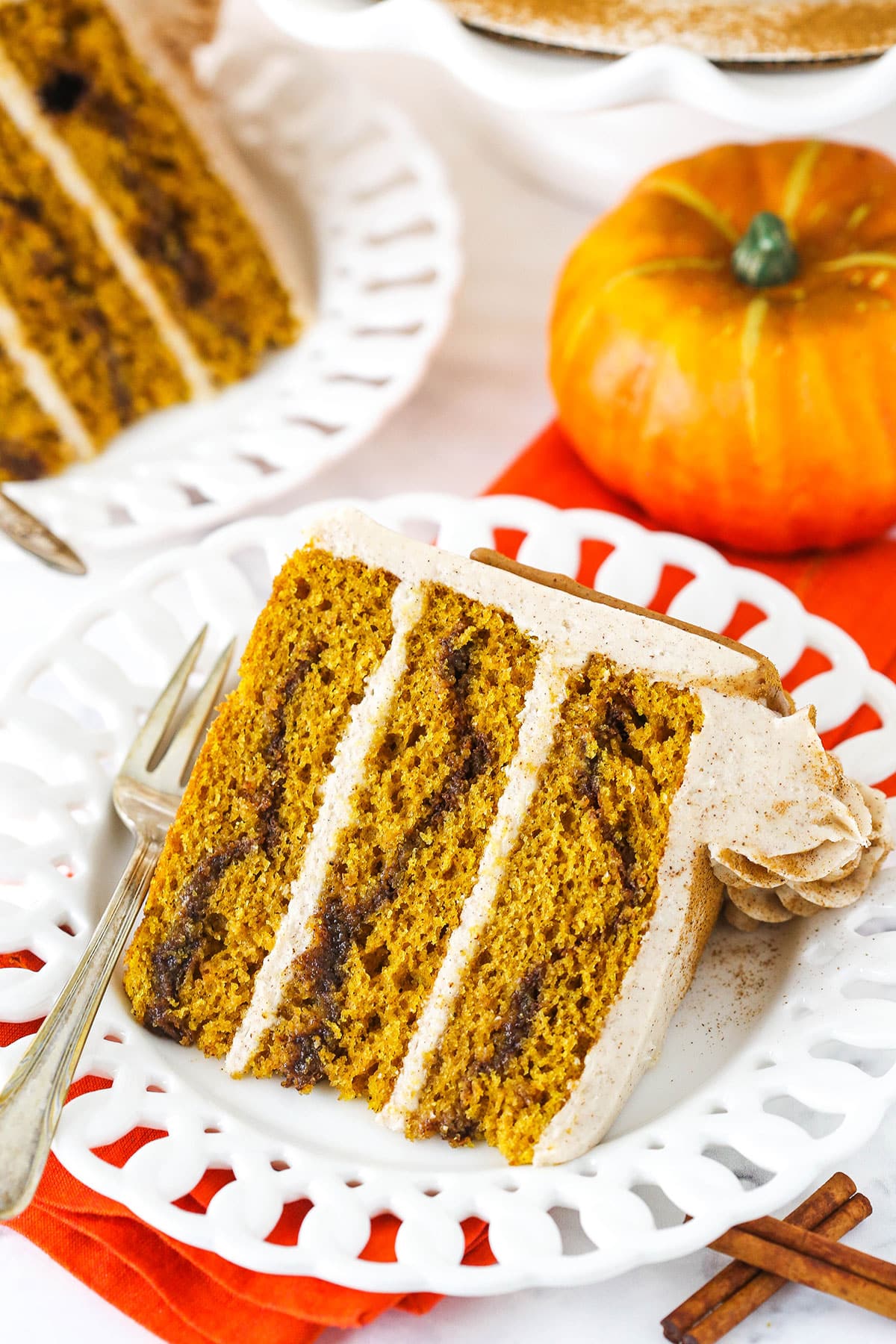 This pumpkin swirl cake is a moist pumpkin cake swirled with cinnamon and sugar in every layer. It's also covered in cream cheese frosting! A slice of Cinnamon Sugar Swirl Pumpkin Layer Cake on it's side on a white plate with a fork.
