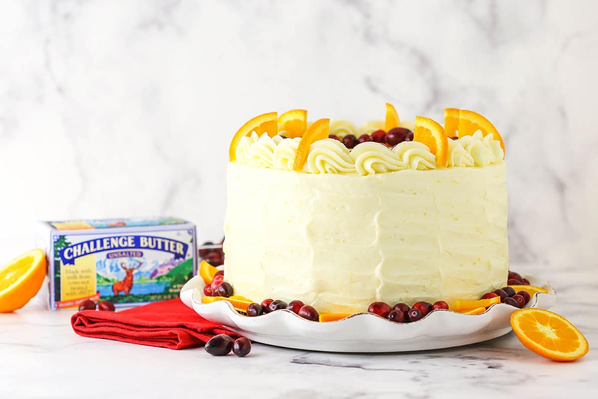 Side view of a full Cranberry Orange Layer Cake on a white platter with butter in background