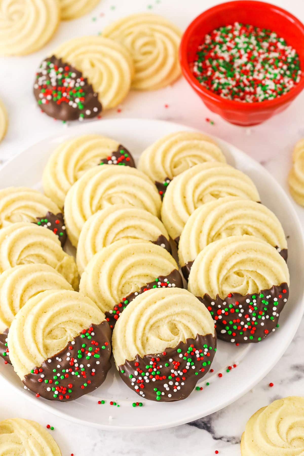 Butter cookies lined up on a plate beside a bowl of sprinkles.
