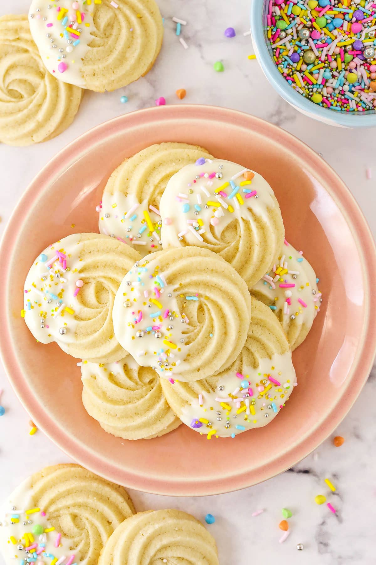 White chocolate butter cookies on a pale pink plate beside a bowl of pastel sprinkles.