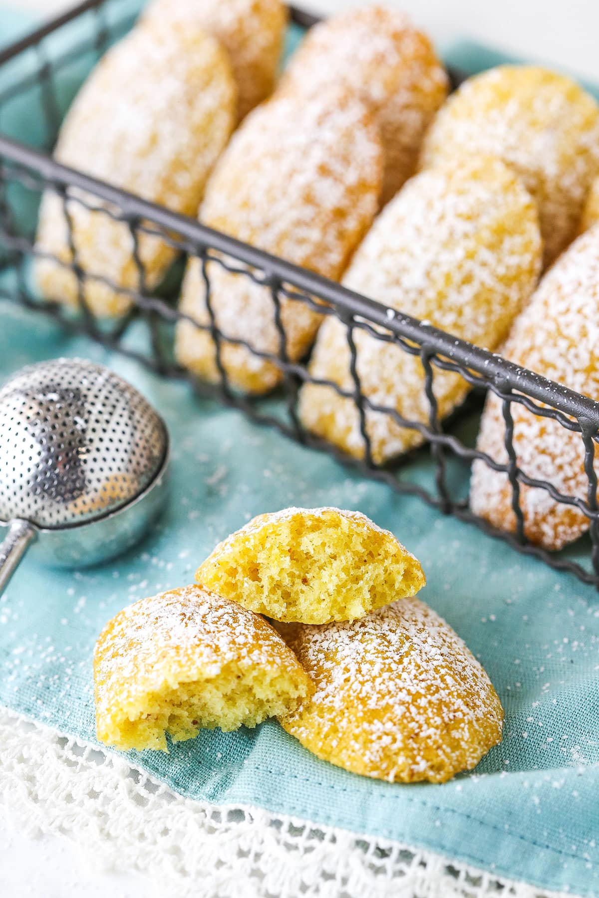 Homemade madeleine cookies are soft, buttery tea cakes dusted with powdered sugar. Make them as a casual treat, or for the holidays! A madeleine tea cake broken in half on a teal napkin with other cookies nearby.