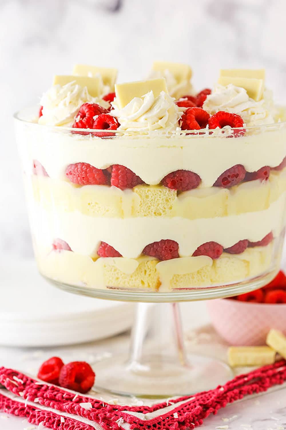 This White Chocolate Raspberry Trifle is a holiday-worthy dessert with layers of fresh raspberries, chocolate ganache, and creamy mousse. Side view of a white chocolate raspberry trifle in a clear trifle dish on top of a red cloth.