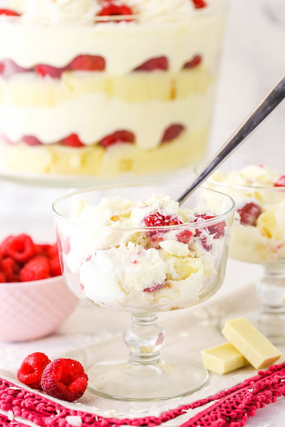 A serving of raspberry trifle in a coup glass with a spoon, with the rest of the trifle in the background.