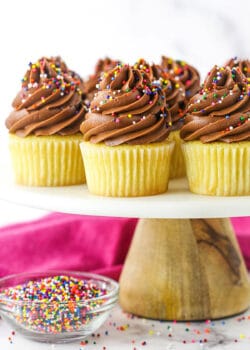 Yellow Cupcakes with Chocolate Frosting on a Cake Stand Next to a Bowl of Sprinkles