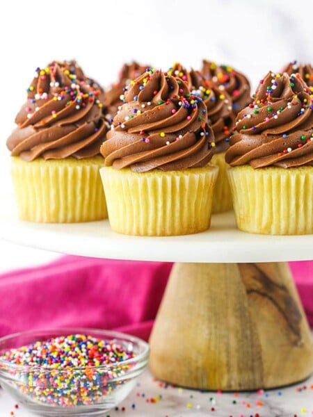Yellow Cupcakes with Chocolate Frosting on a Cake Stand Next to a Bowl of Sprinkles