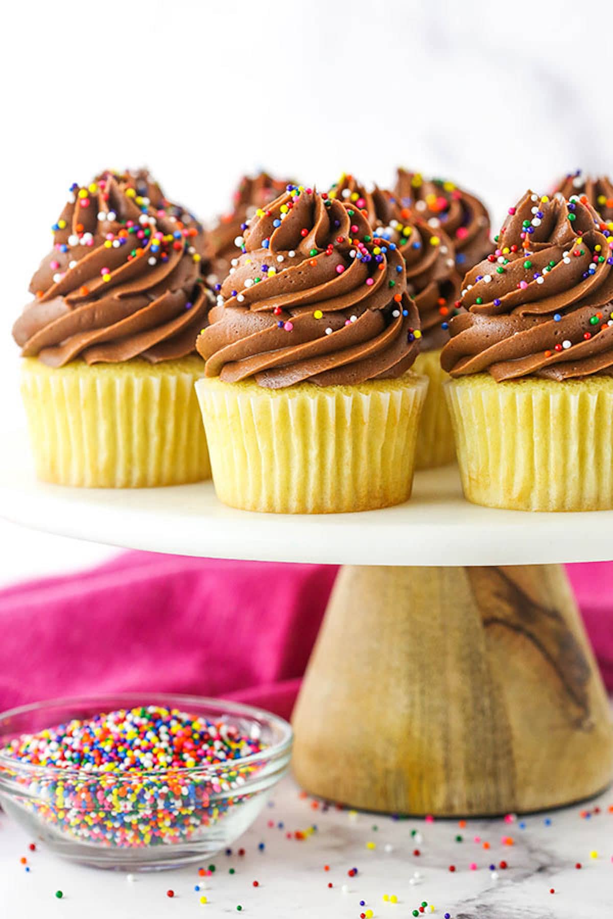 Side view of yellow cupcakes with chocolate frosting and sprinkles on a cake stand next to a bowl of sprinkles. 