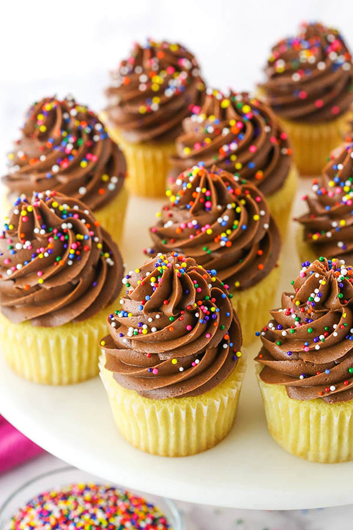 A platter full of yellow cupcakes with chocolate frosting and sprinkles.