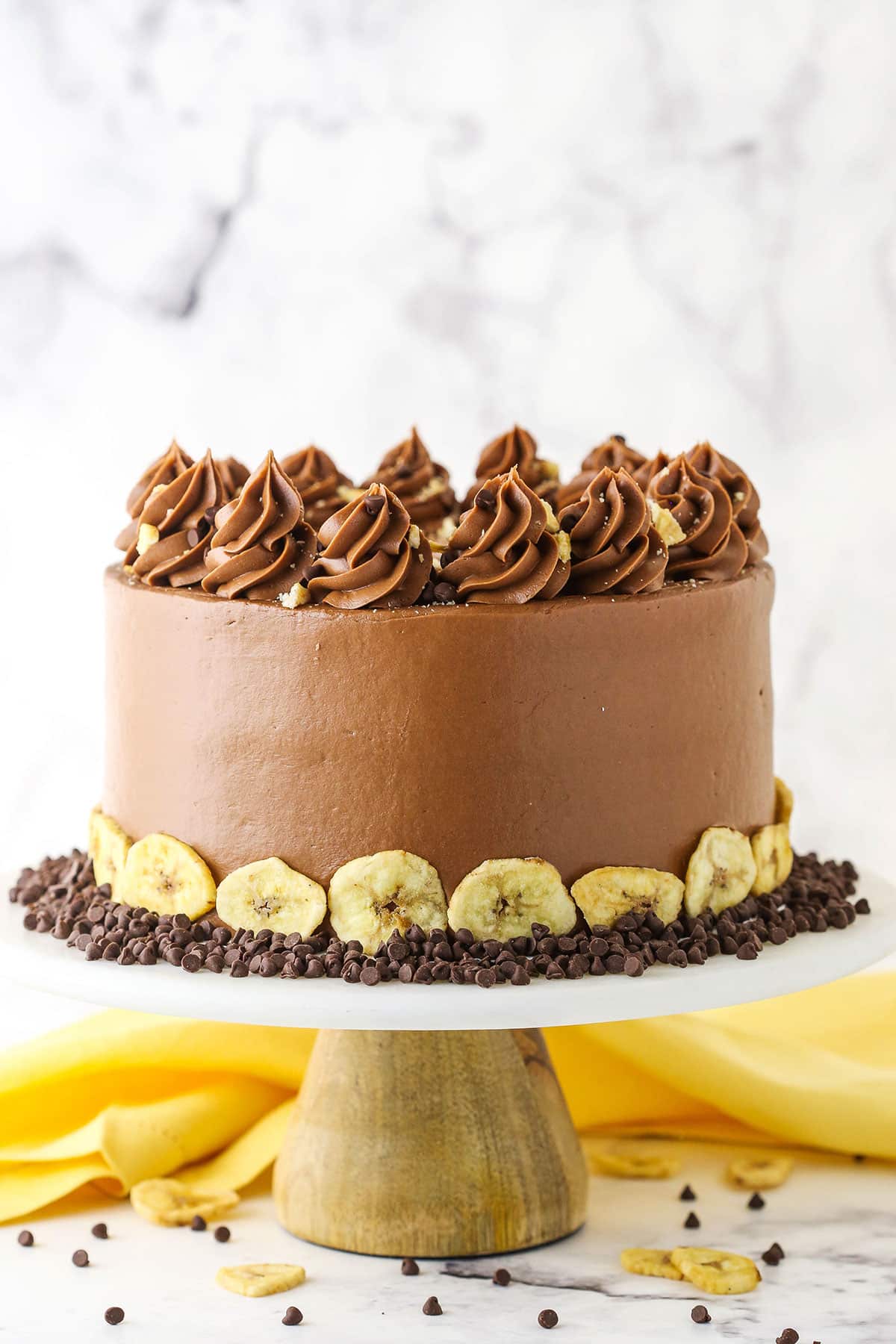 Side view of a full banana chocolate chip cake, topped with chocolate swirls, chocolate chips and banana chips on a wood and marble cake stand