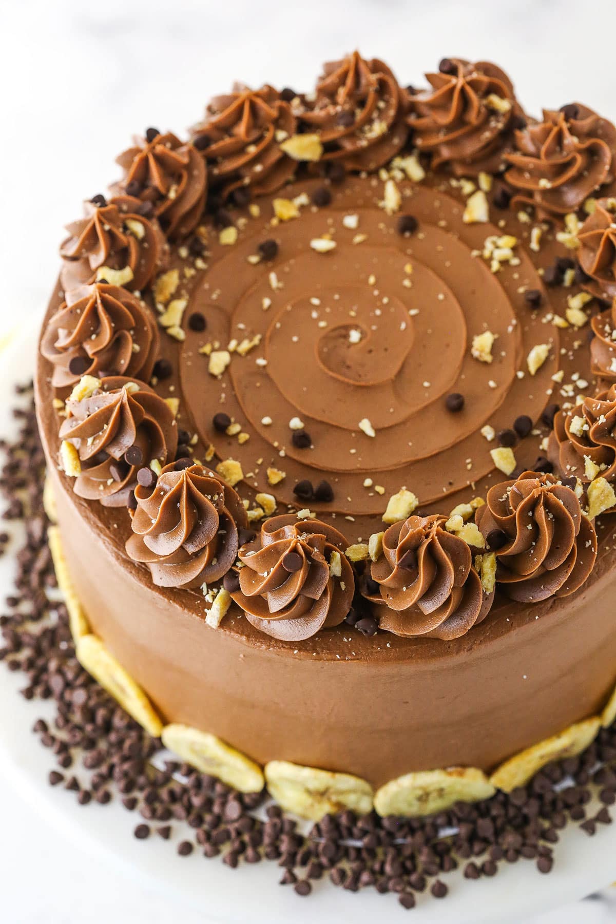 Overhead view of banana chocolate cake with chocolate frosting swirls and bits of crushed banana chips and chocolate chips on top.