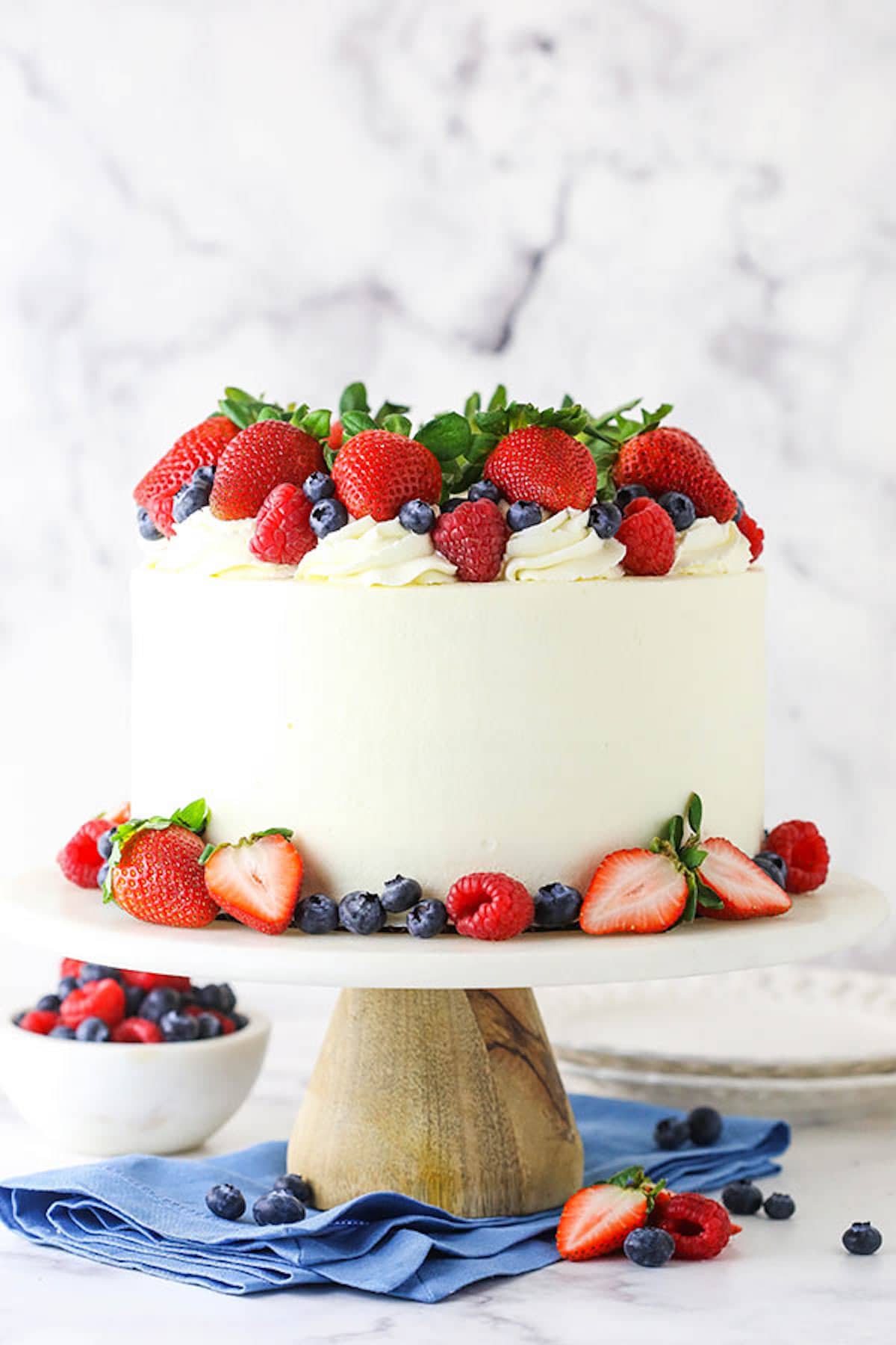 A Berry Chantilly Layer Cake on a Wooden Cake Stand Next to a Bowl of Berries and a Stack of Paper Plates