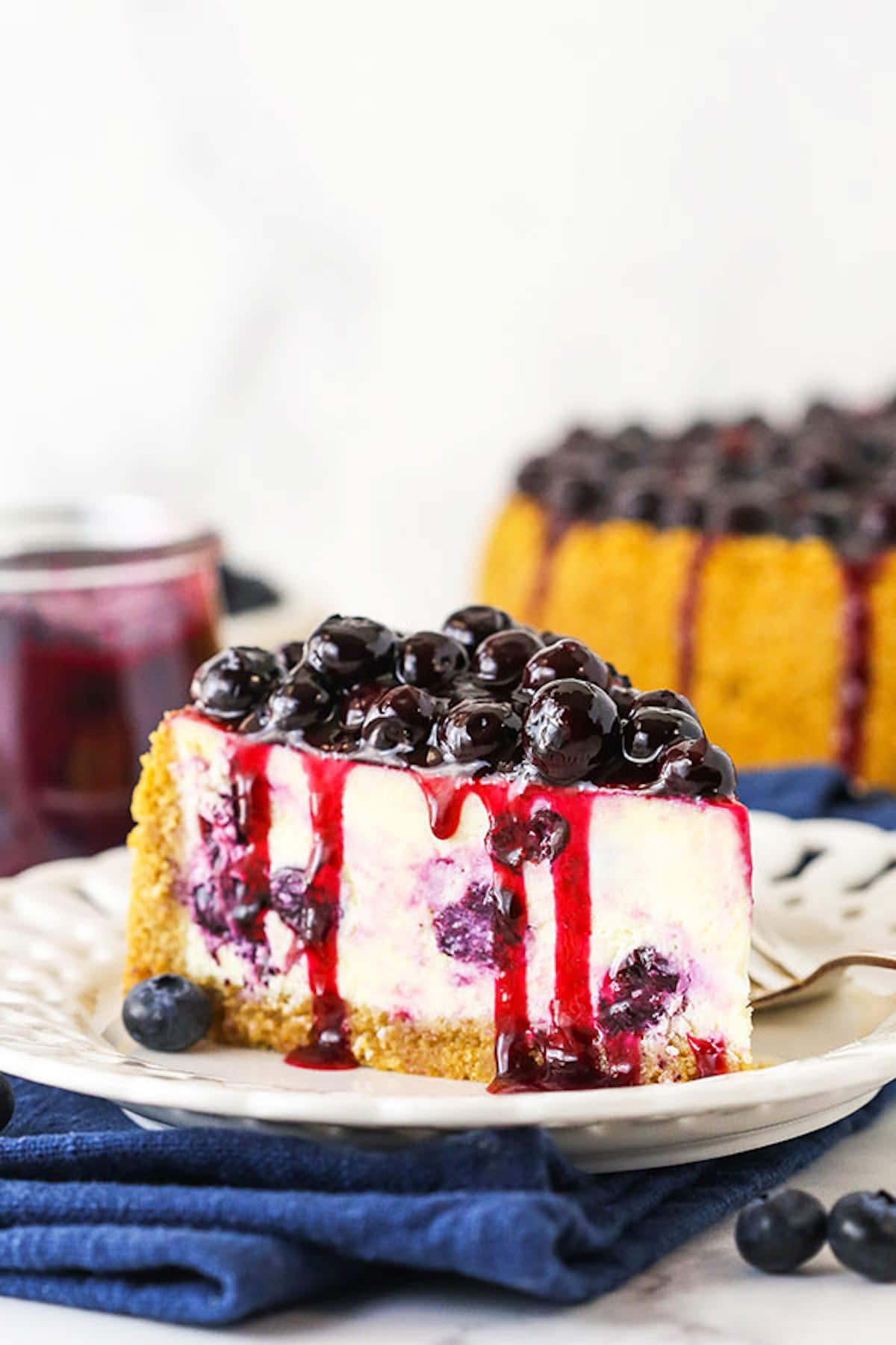 A Piece of Blueberry Cheesecake with a Bowl of Sauce and the Rest of the Cheesecake in the Background