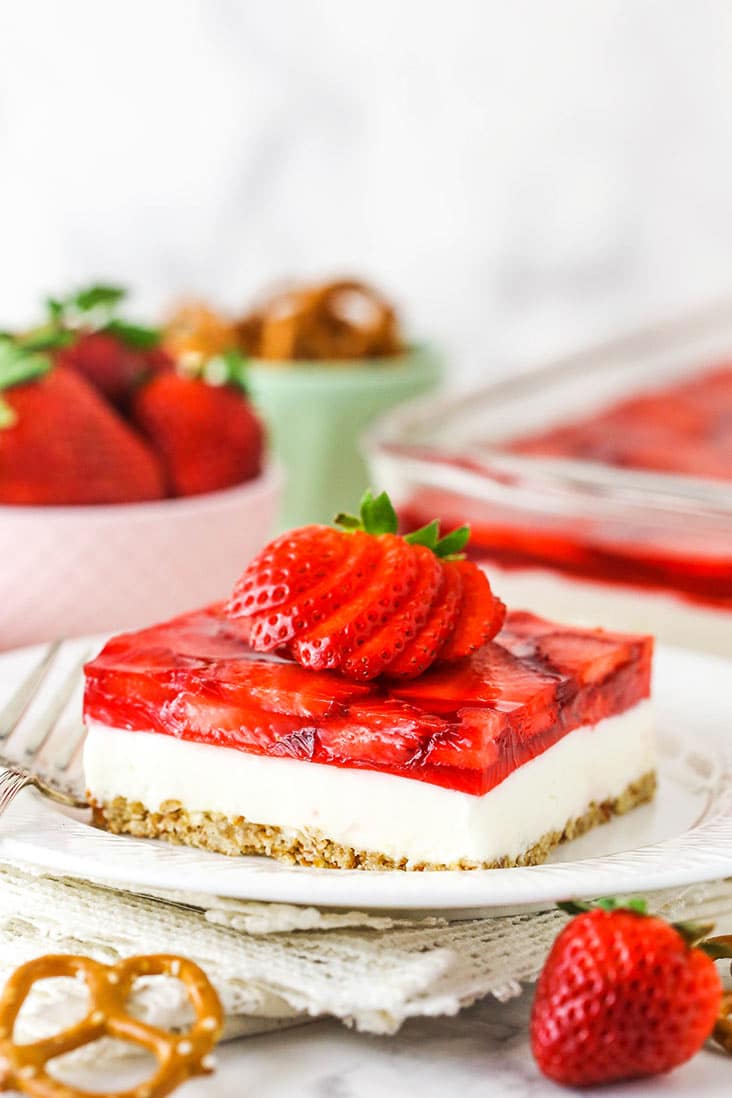 A square slice of strawberry pretzel salad on a white plate, with a strawberry and pretzel in front of the plate.
