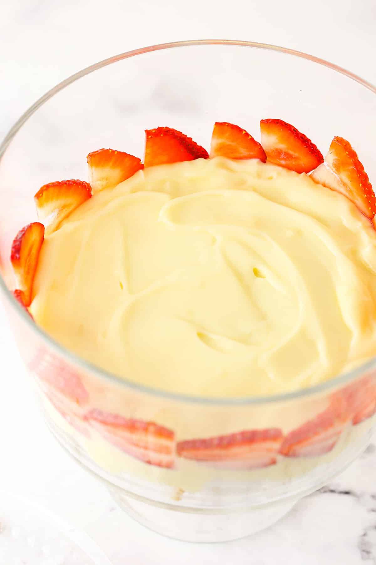 This trifle recipe is the best summer dessert! It's filled with layers of angel food cake, pudding, strawberries, blueberries & raspberries. Overhead view of strawberries being added on top of trifle pudding layer.