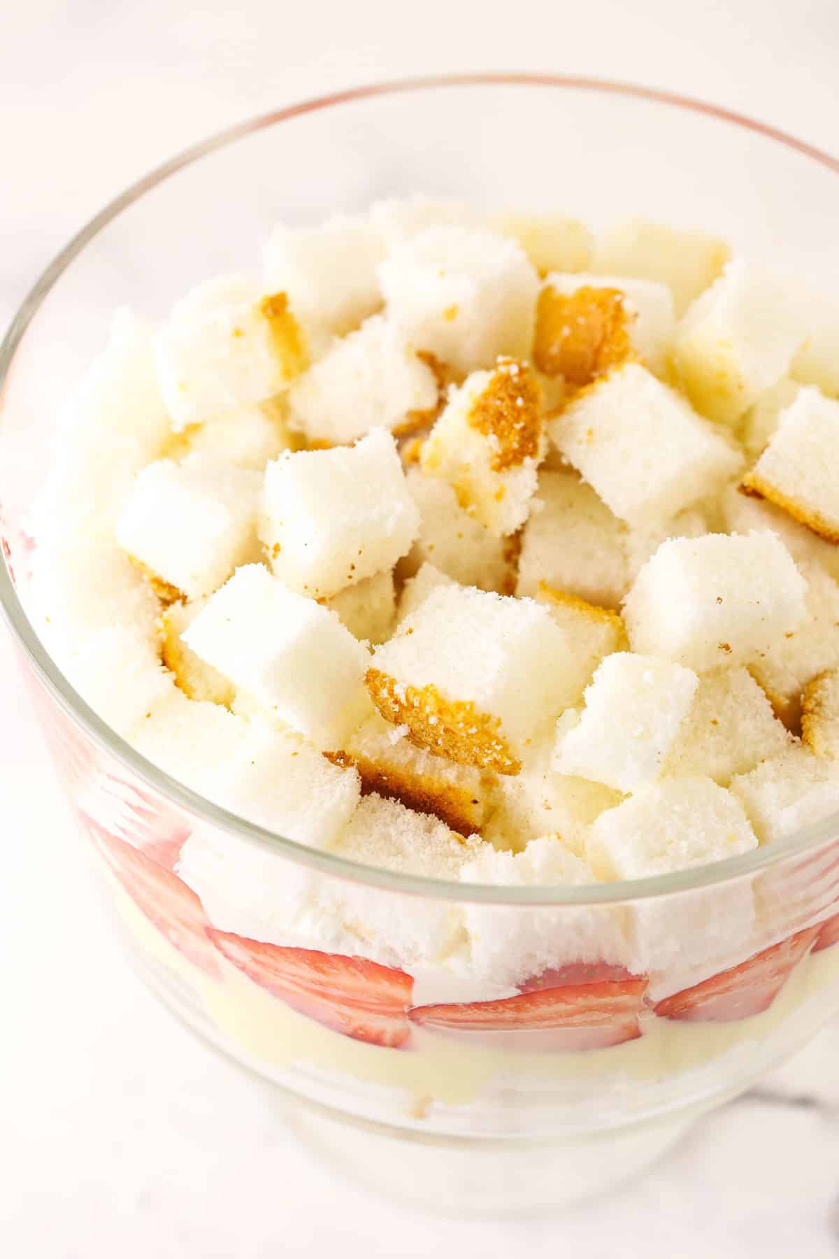 Overhead view of angel food cake cut into cubes being added on top of trifle whipped cream layer.