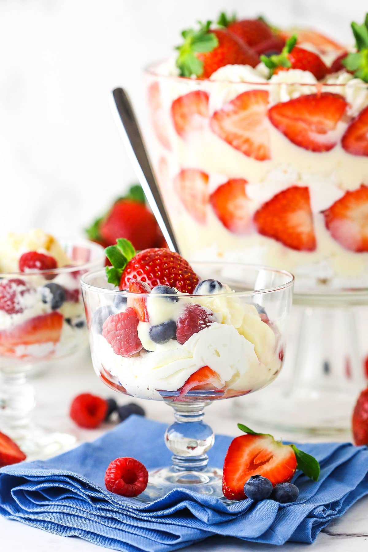 This trifle recipe is the best summer dessert! It's filled with layers of angel food cake, pudding, strawberries, blueberries & raspberries. A scoop of trifle in a mini trifle serving dish on a blue napkin