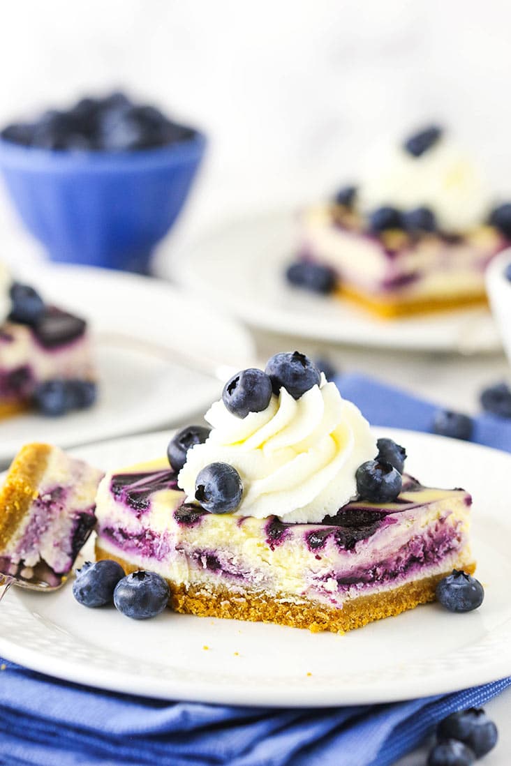 A slice of blueberry cheesecake topped with whipped cream and blueberries, with a bite missing