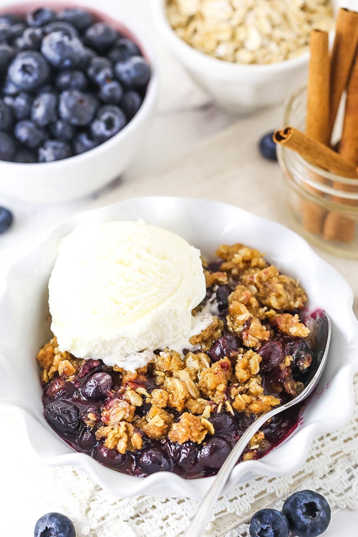 blueberry crisp with a scoop of ice cream in white bowl with blueberries, oats and cinnamon sticks in background