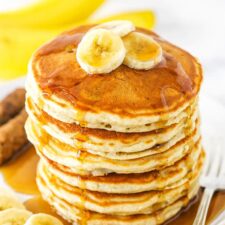 Banana pancakes stacked high on a plate with syrup and banana slices