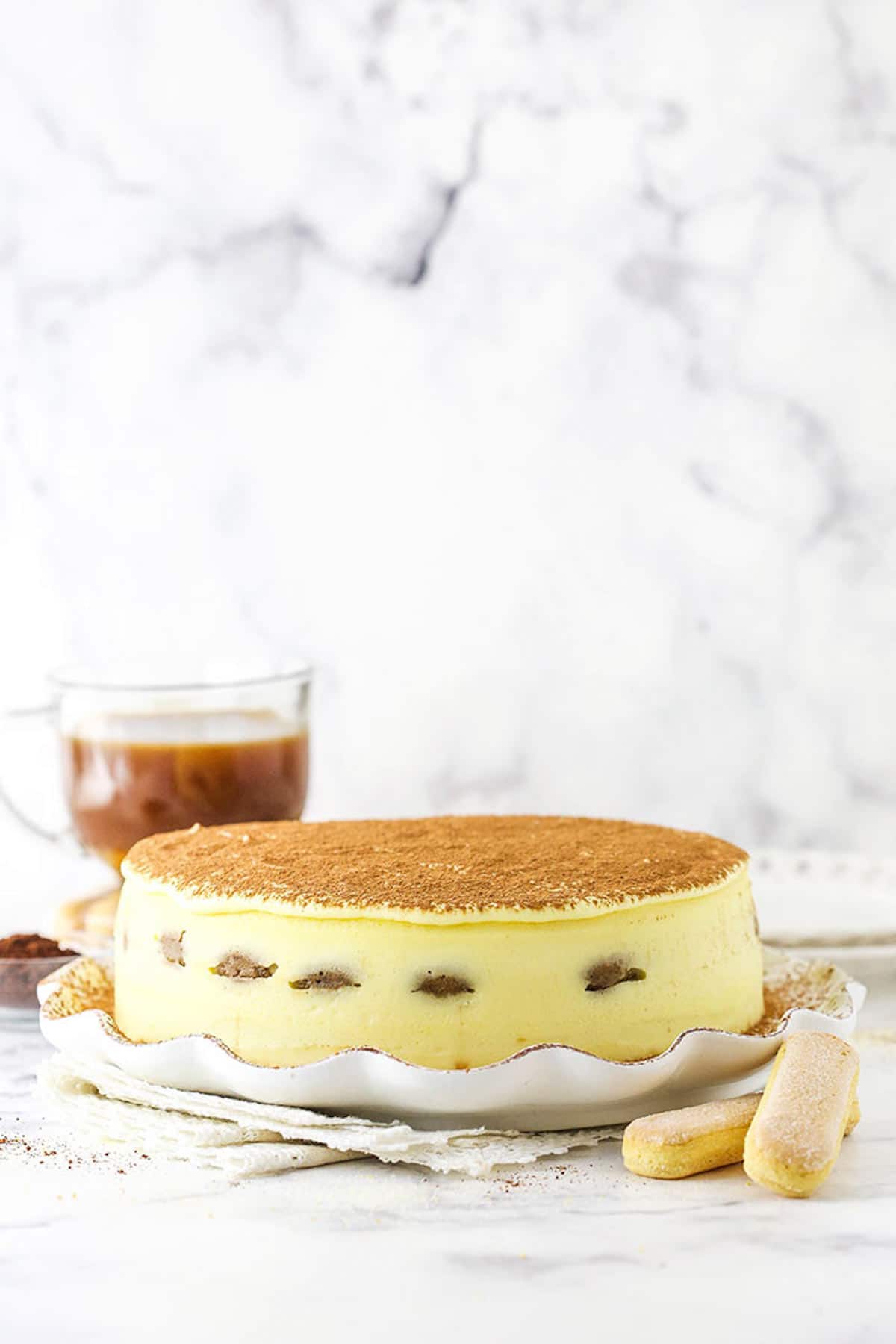 A Tiramisu Cheesecake on a Black and White Marble Countertop Beside Two Ladyfingers