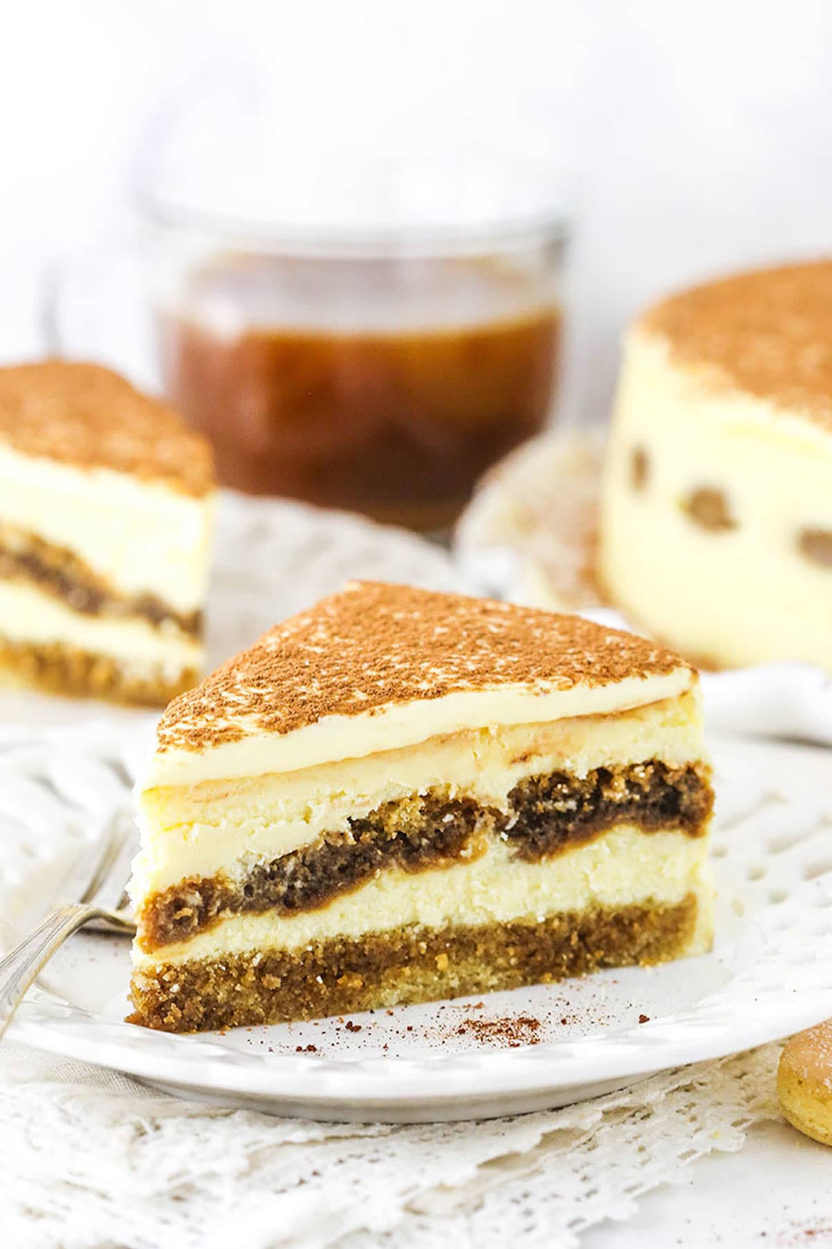 A Piece of Tiramisu Cheesecake on a Plate with a Second Slice and the Full Cake in the Background