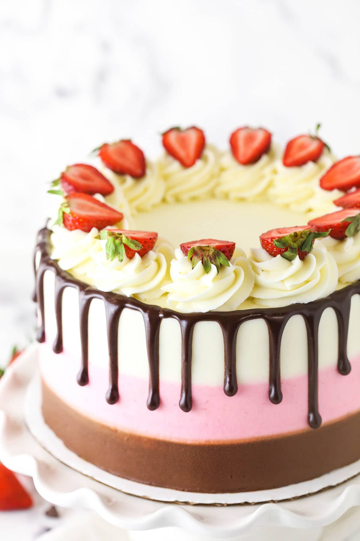 A neapolitan cake decorated with vanilla buttercream swirls, strawberry slices and drips of chocolate ganache