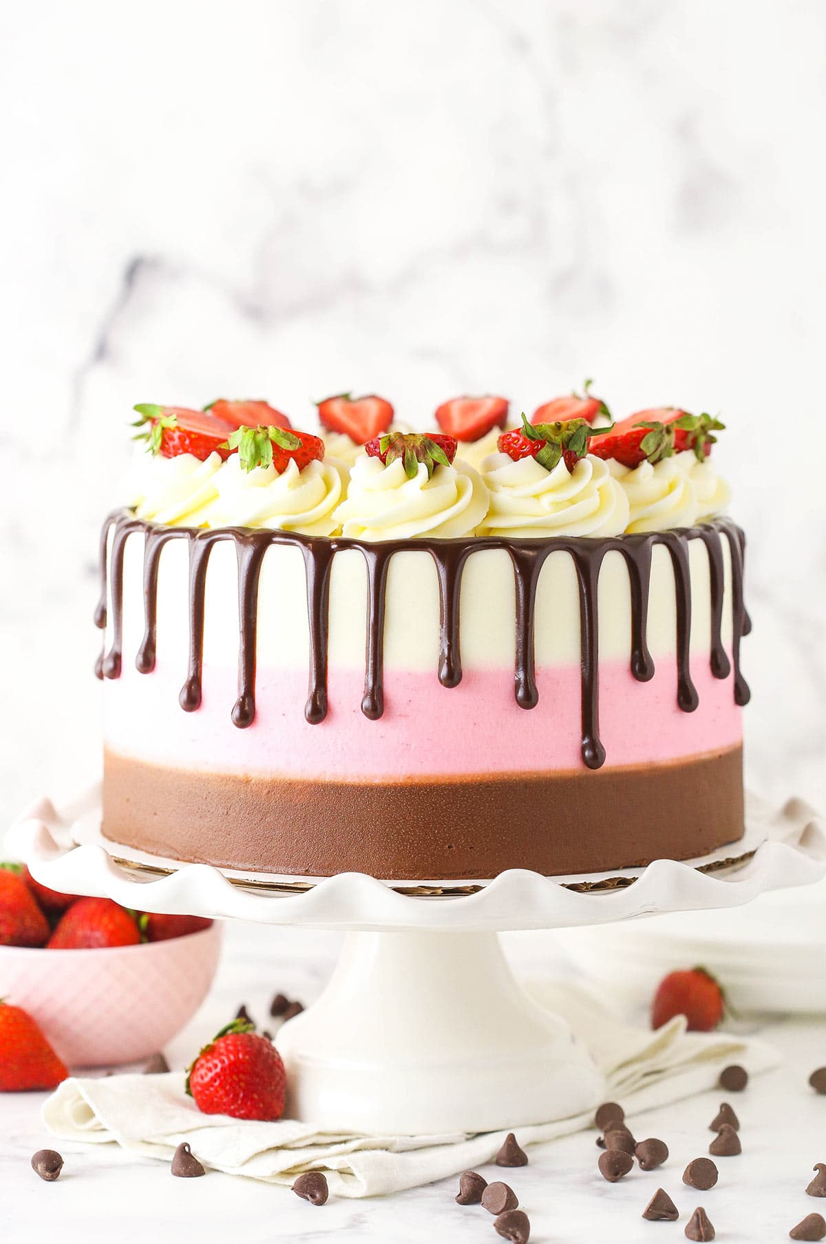 A neapolitan cake sitting on a white cake stand with strawberries and chocolate chips on the counter below