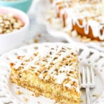 A piece of toffee almond coffee cake on a plate with the full cake and bowls of almonds and toffee in the background