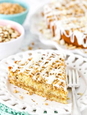 A piece of toffee almond coffee cake on a plate with the full cake and bowls of almonds and toffee in the background