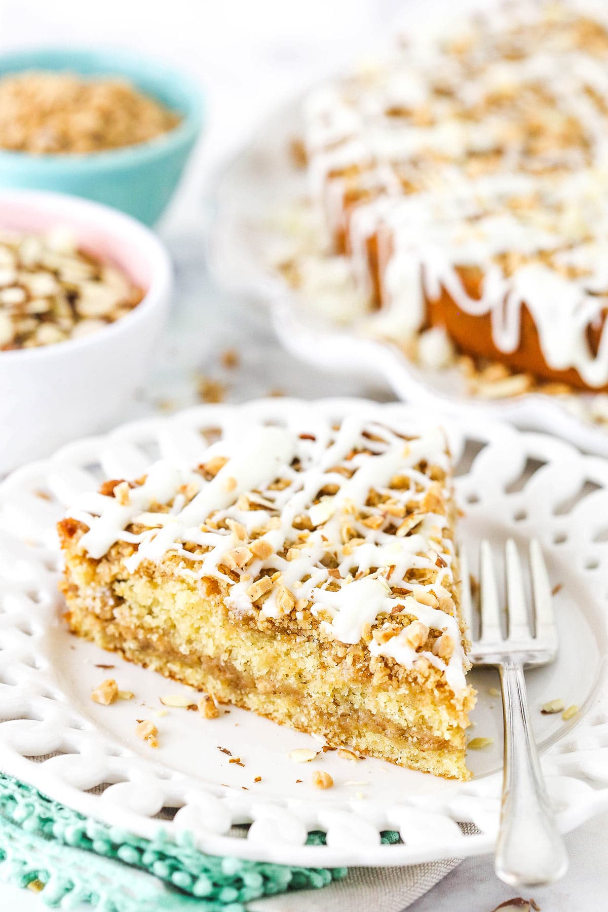 A piece of toffee almond coffee cake on a plate with the full cake and bowls of almonds and toffee in the background