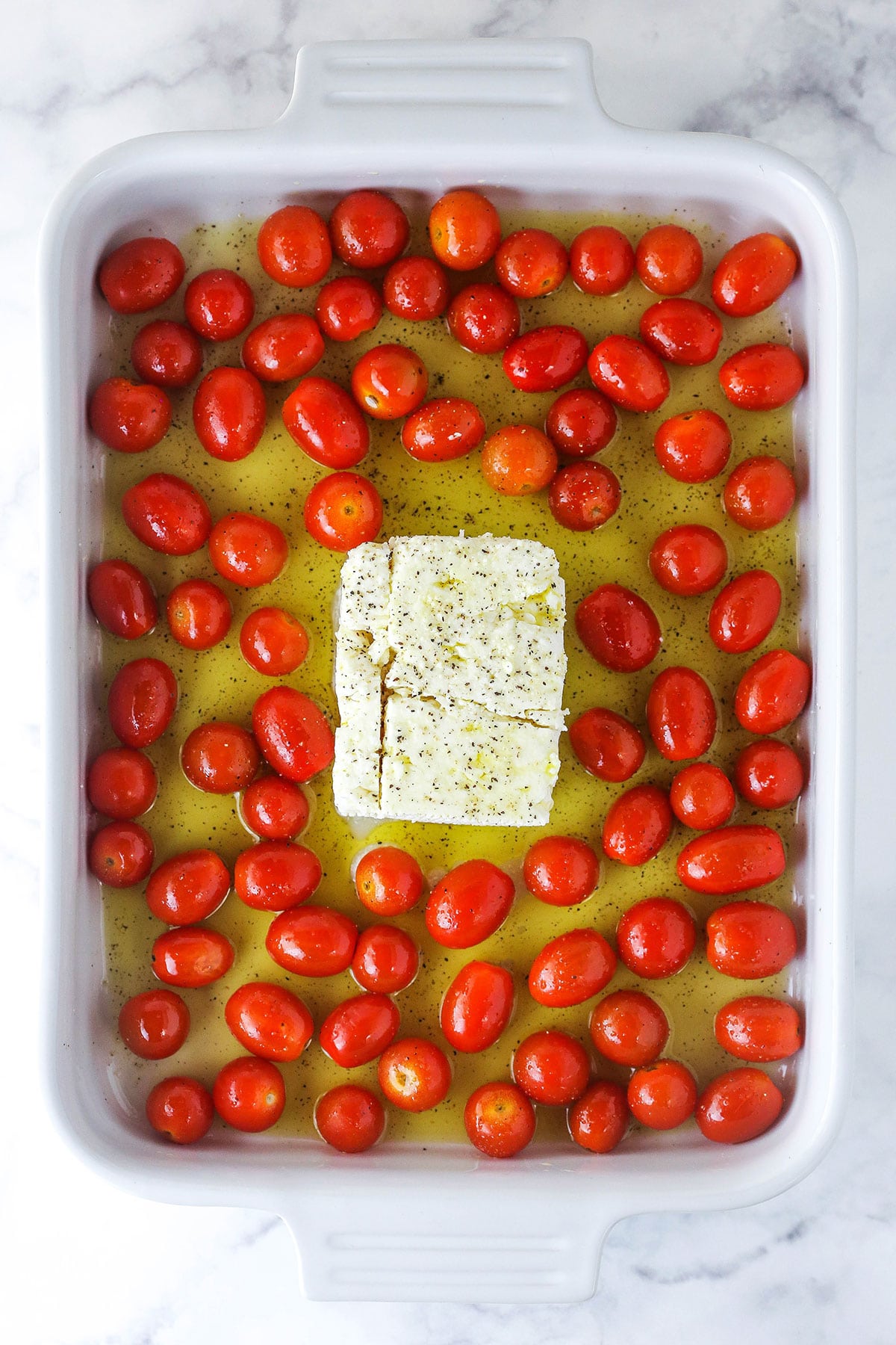 Extra virgin olive oil, salt, pepper and cherry tomatoes surrounding a block of feta cheese in a baking dish