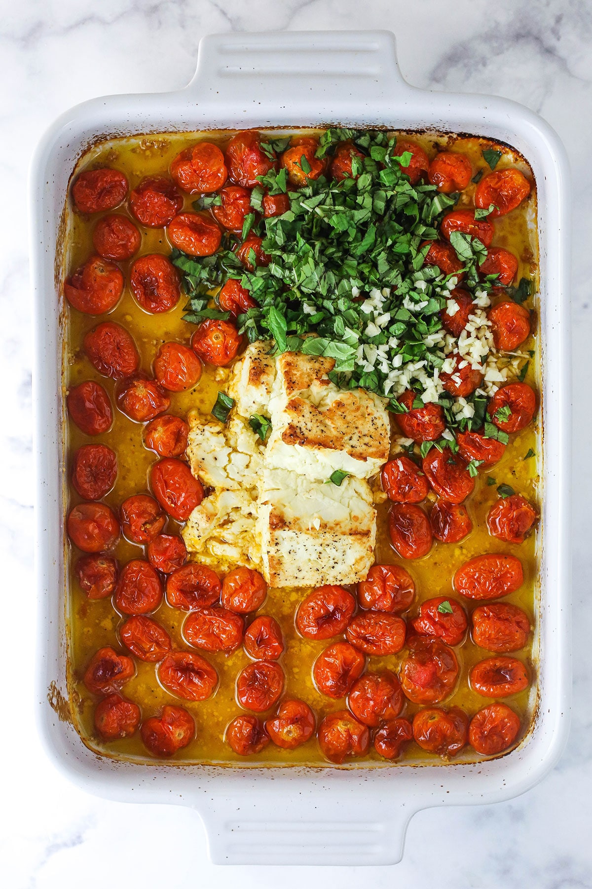 A casserole dish full of baked feta, oil and cherry tomatoes with chopped fresh basil and minced garlic poured on top