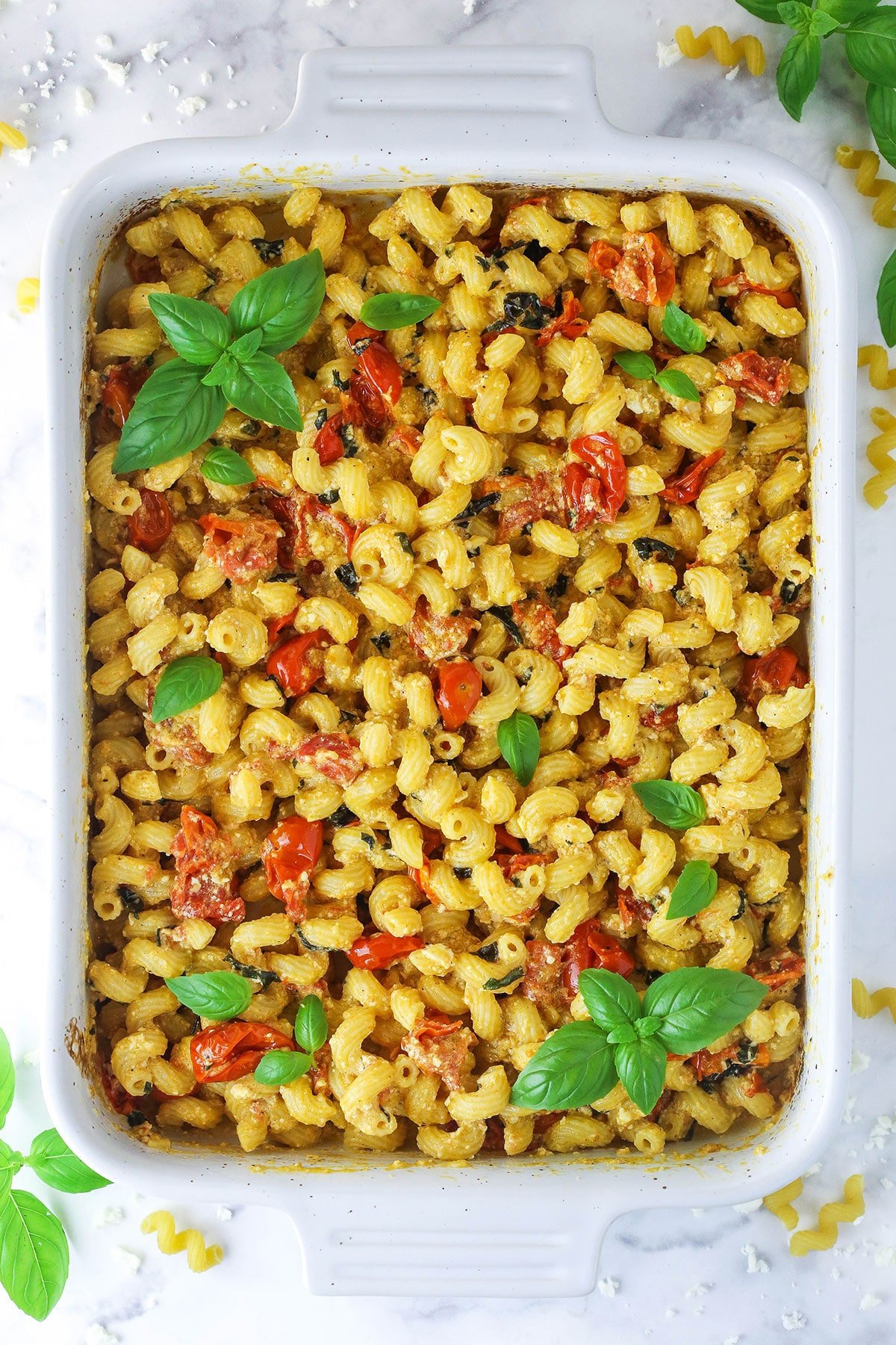 A baking dish full of cheesy pasta bake on a countertop with dry noodles and basil leaves