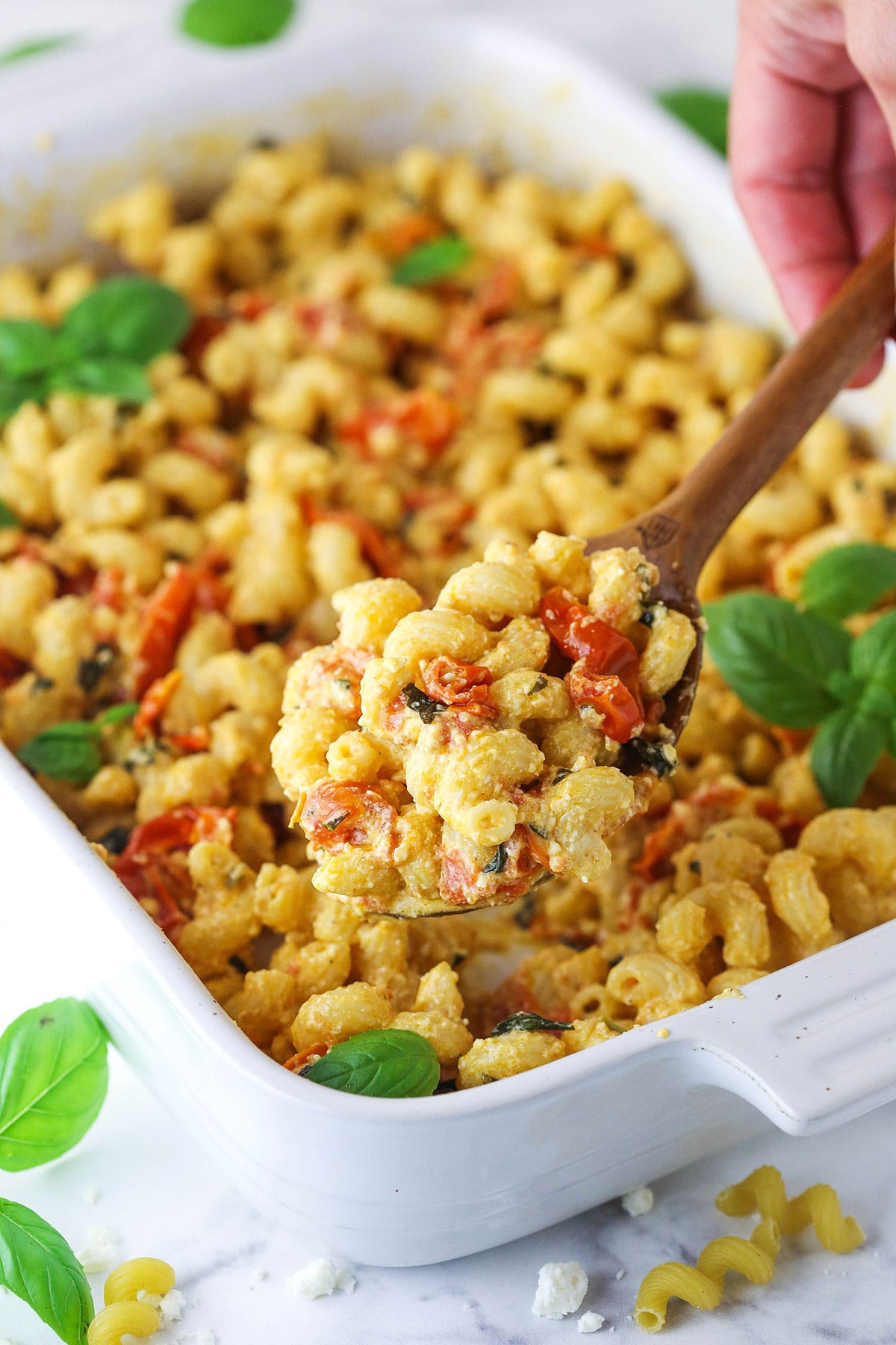 A wooden spoon scooping out a spoonful of baked feta pasta from a white baking dish