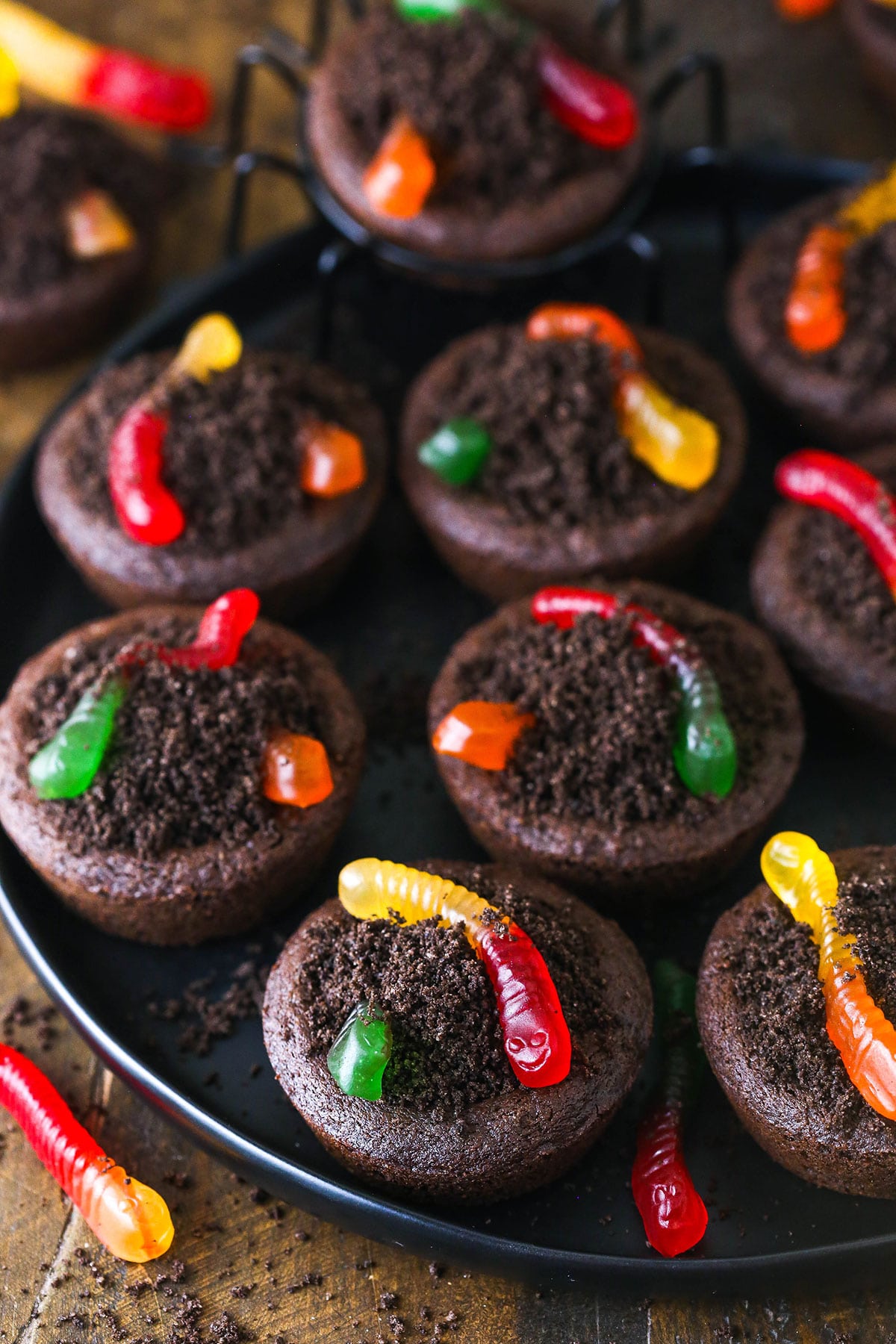 A round, black platter full of Halloween cookies topped with crushed Oreo cookies and two gummy worms