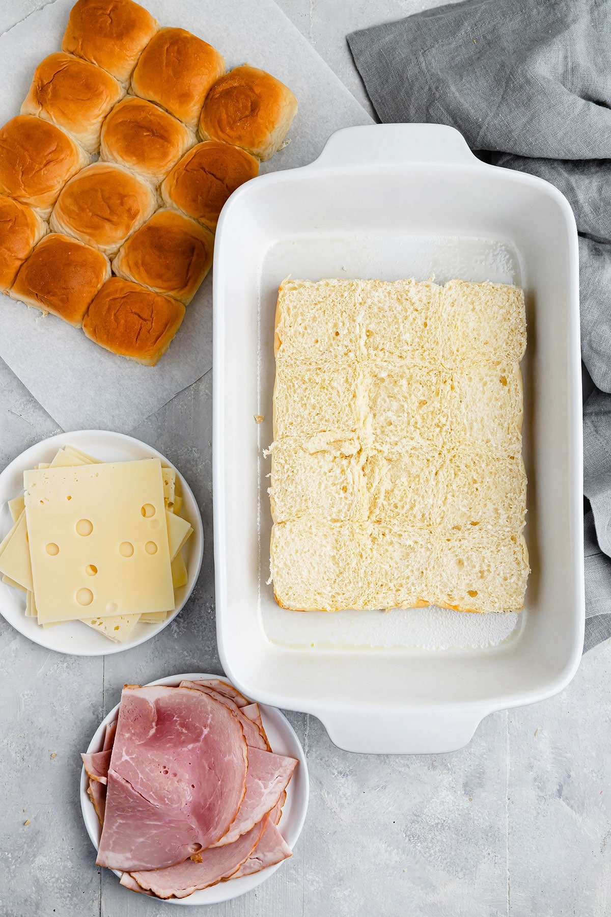 Halved Hawaiian rolls, Swiss cheese and ham on a gray countertop beside a kitchen towel