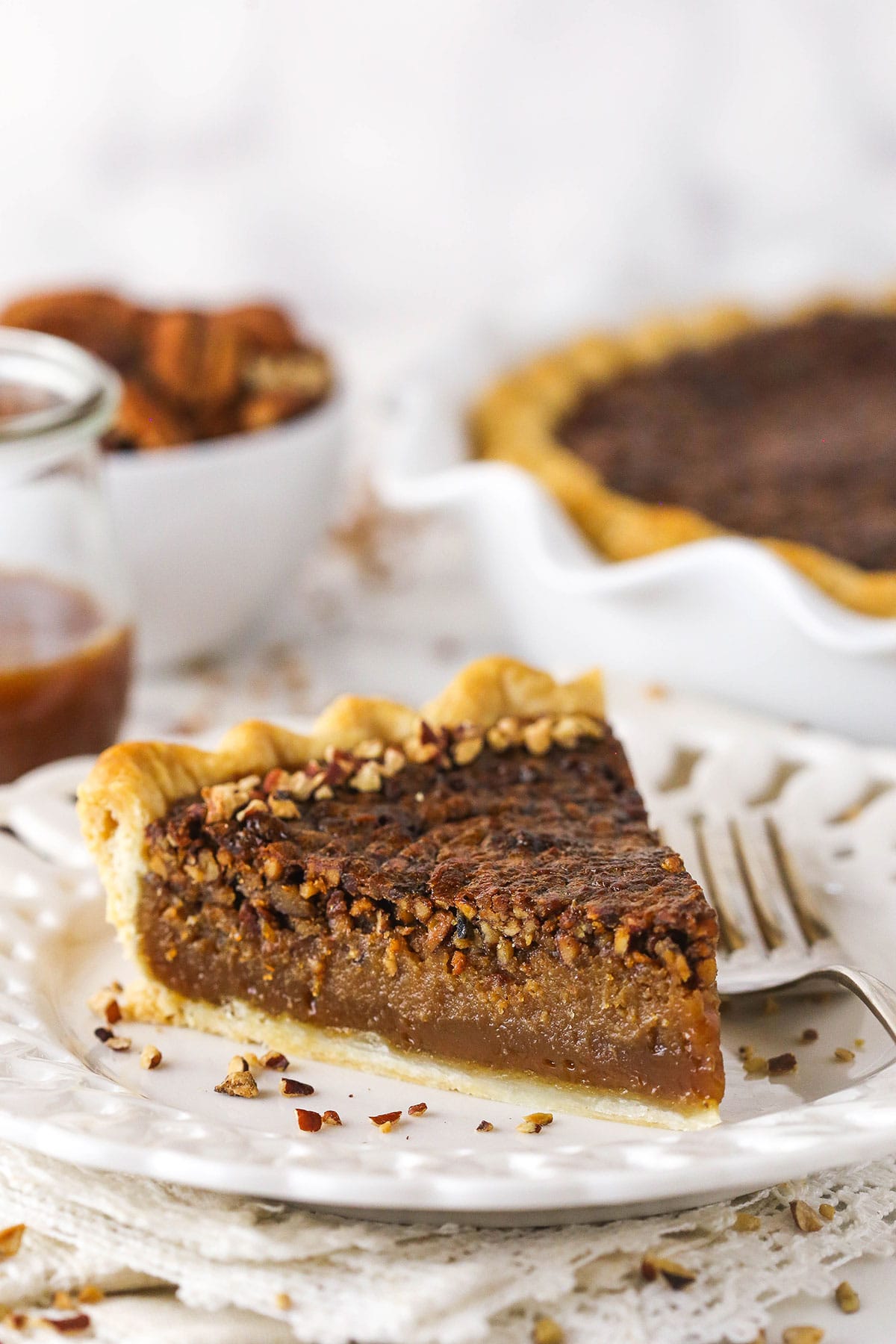 One piece of caramel pecan pie on a plate with a bowl of pecans and a full pie behind it