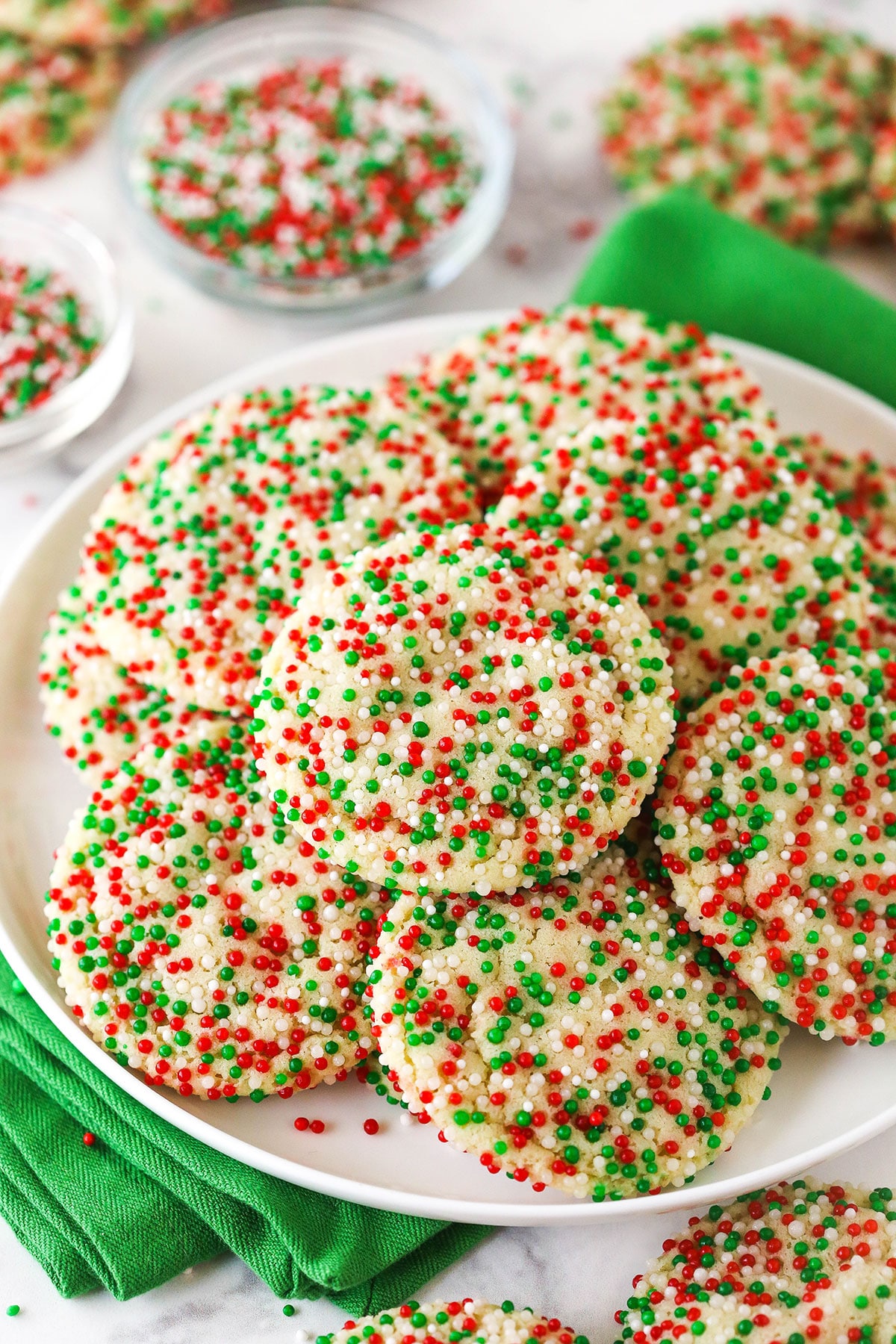 A plate of Christmas sugar cookies on top of a green kitchen towel with a bowl of sprinkles behind it