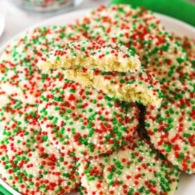 Christmas sprinkle cookies piled onto a white plate with one broken in half to reveal the soft and fluffy interior