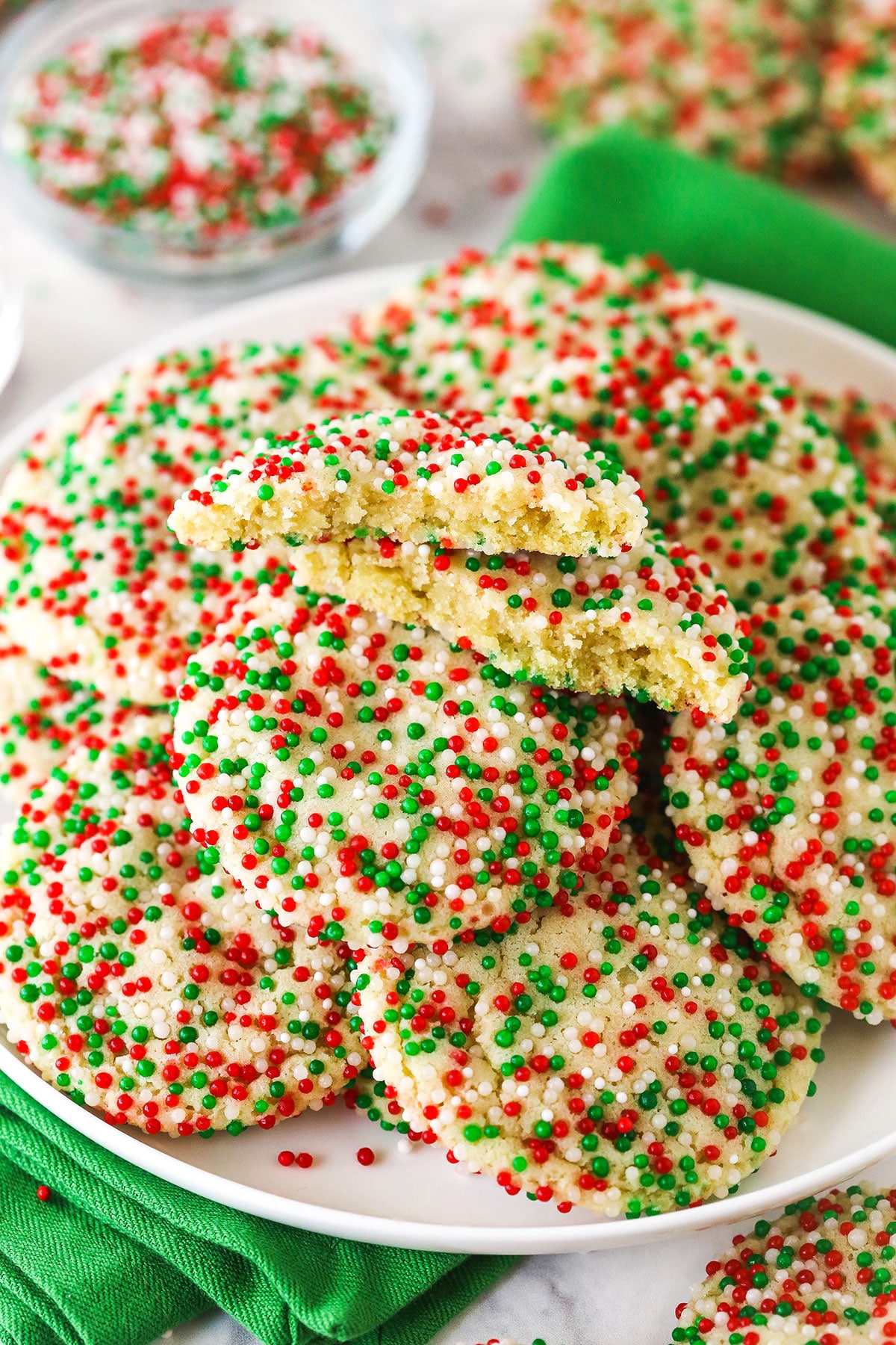 Christmas sprinkle cookies piled onto a white plate with one broken in half to reveal the soft and fluffy interior