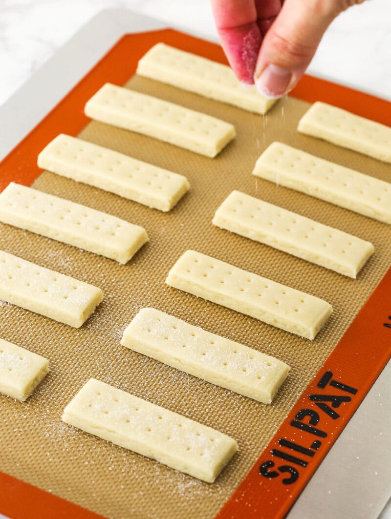 Sugar being sprinkled over twelve docked sections of dough on a lined baking sheet.