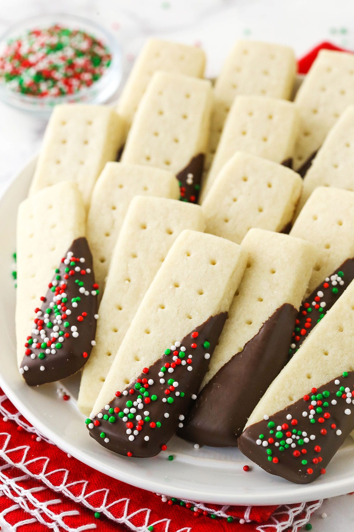 A serving platter full of shortbread cookies with a small bowl of Christmas sprinkles in the background.