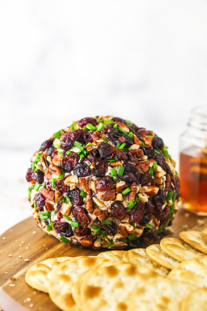 A homemade cheeseball on top of a wooden cutting board beside chips and a jar of honey