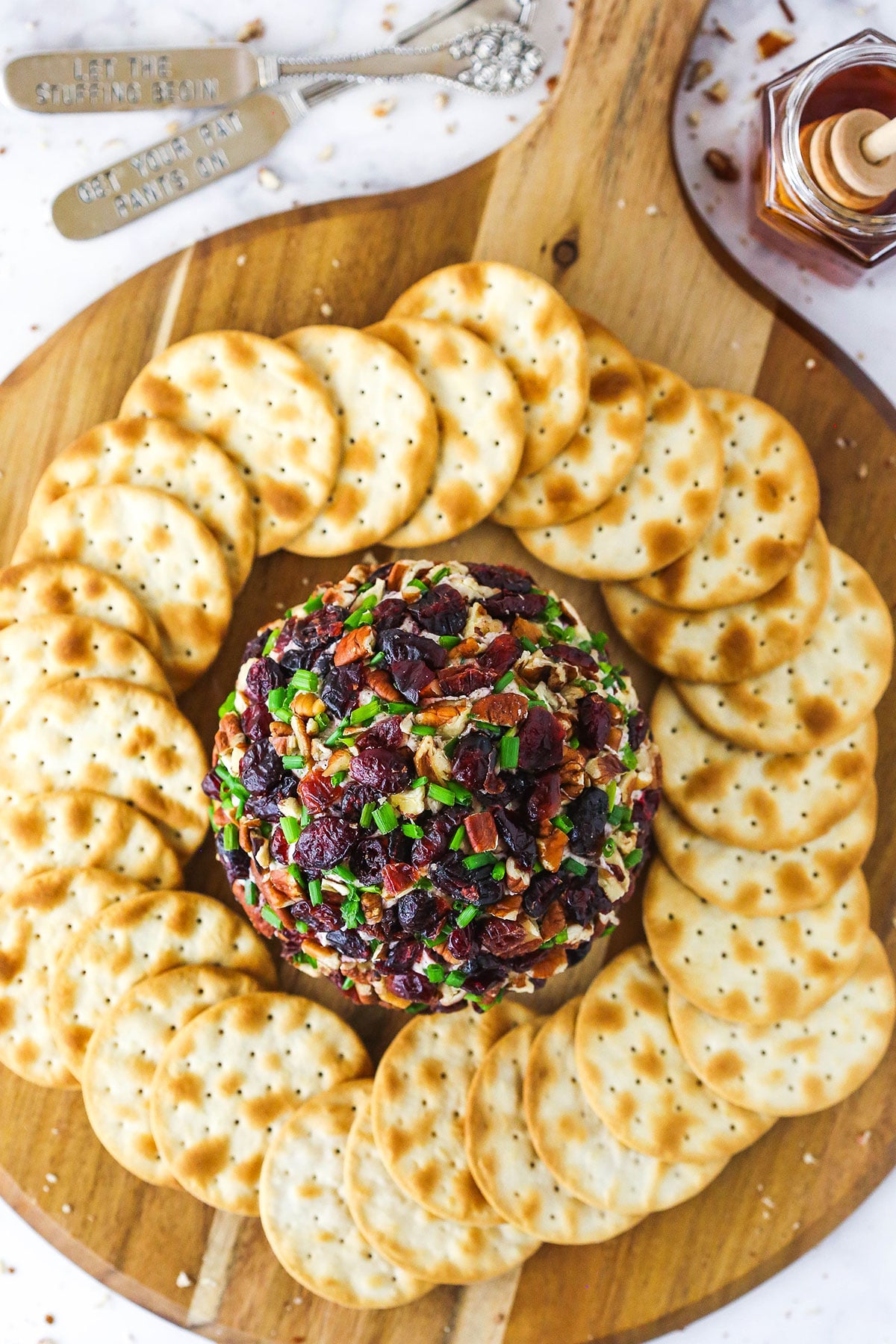A cranberry pecan cheeseball surrounded by crackers with a small dish of honey beside the appetizer