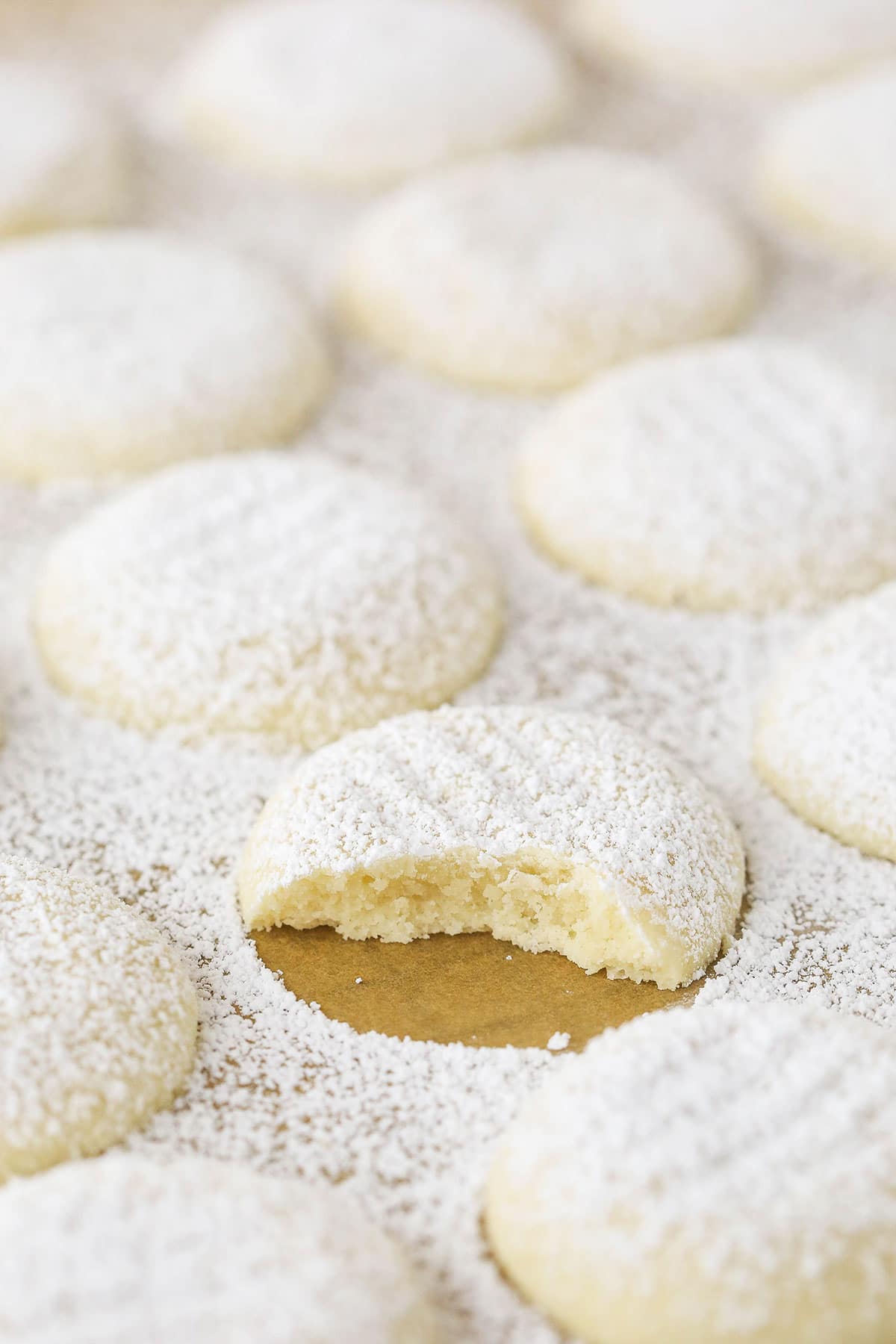 A uniform arrangement of powdered sugar coated vanilla butter cookies with one missing a bite.