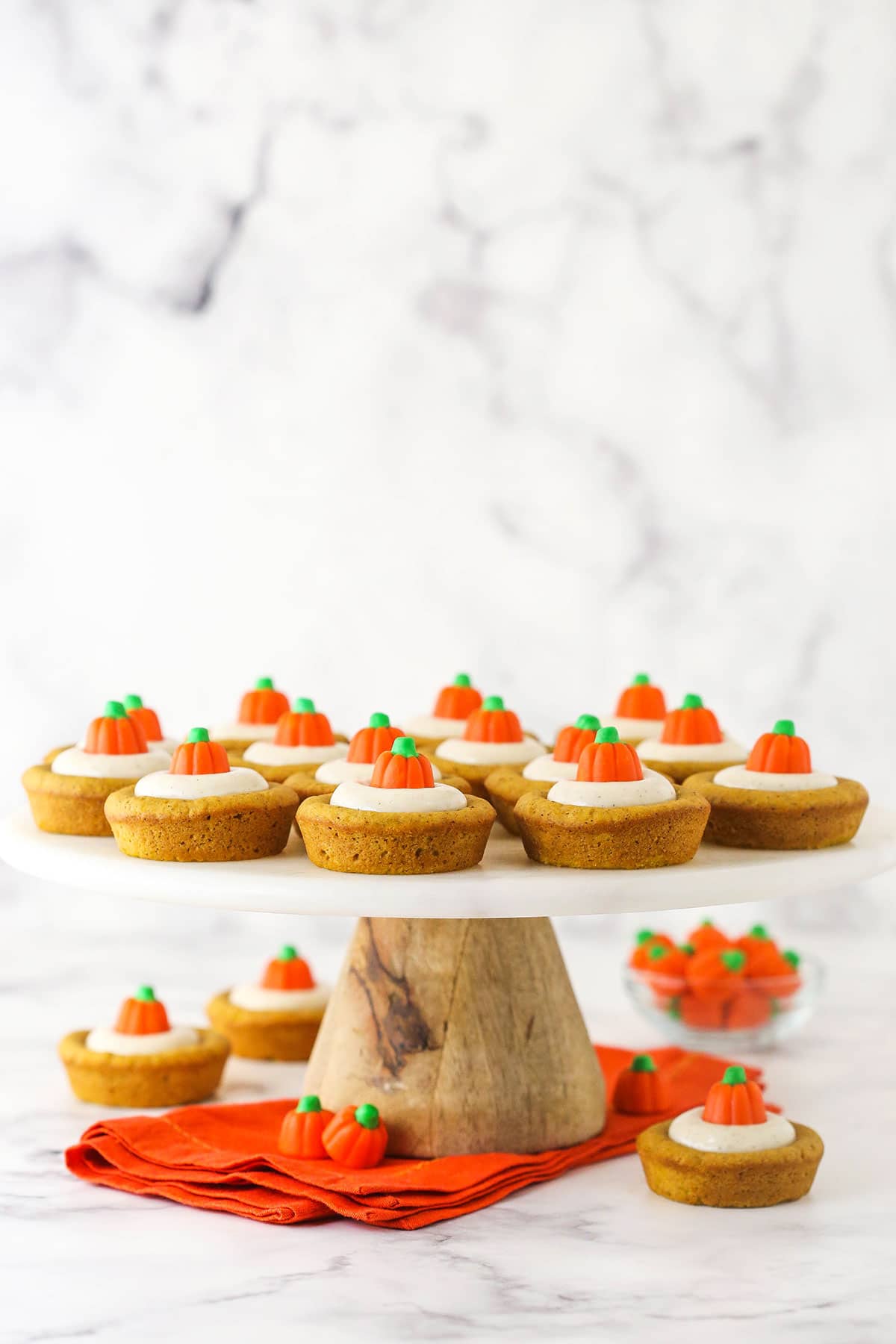 A marble counter holding a cake stand displaying a batch of pumpkin cookies with more cookies and candy pumpkins underneath it