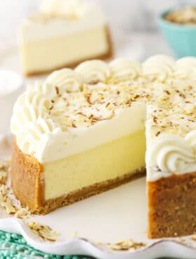 An almond mousse cheesecake on a serving platter with one slice removed and featured in the background