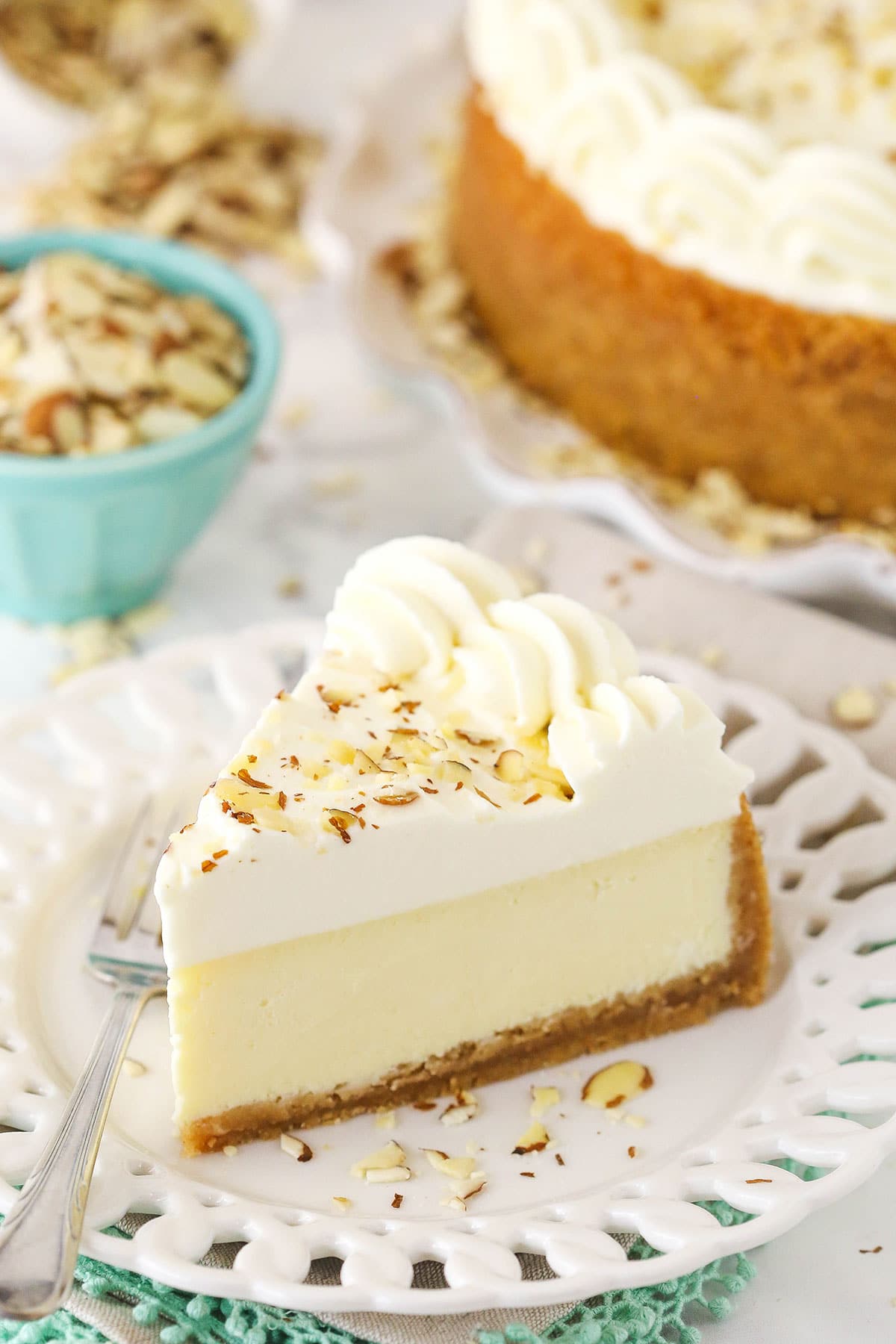 A piece of almond mousse cheesecake on a white plate with the full cheesecake in the background