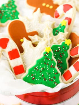 Sugar cookies shaped like snowflakes, candy canes and Christmas trees inside of a metal tin on a kitchen countertop